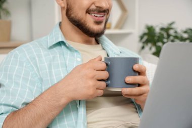 Seramik bardaklı ve dizüstü bilgisayarlı genç adam evde oturuyor, yakın plan.