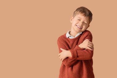 Sıcak süveter giyen mutlu küçük çocuk bej arka planda kendine sarılıyor.