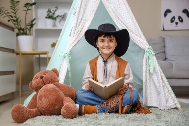 Kovboy gibi giyinmiş, evinde oyun çadırında macera kitabı okuyan tatlı bir çocuk.