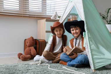 Kovboy gibi giyinmiş şirin çocuklar evdeki çadırda macera kitabı okuyorlar.