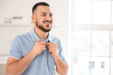 Genç adam evde mavi polo gömleğini ilikliyor.