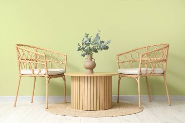 Hasır sandalyeler ve okaliptüs dalları olan kahve masası, yeşil duvarın yanındaki vazoda.