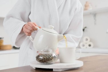 Bornozlu genç kadın çaydanlıktan fincana tatlı çay dolduruyor, yakın plan.
