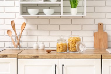 Mutfaktaki beyaz tuğla duvarın yanındaki masada çiğ makarna kabuğu olan kavanozlar.