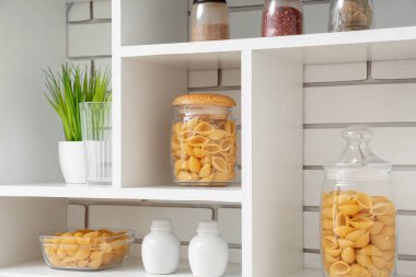 Mutfaktaki beyaz tuğla duvarın yanında raflarda çiğ makarna kabukları olan kavanozlar ve kâse.