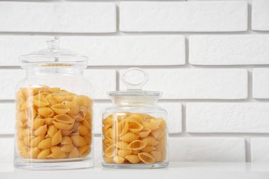 Mutfaktaki beyaz tuğla duvarın yanında rafta çiğ makarna kabukları olan kavanozlar.