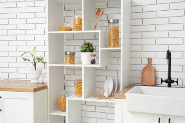 Mutfaktaki beyaz tuğla duvarın yanında raflarda çiğ makarna kabukları olan kavanozlar ve kâse.