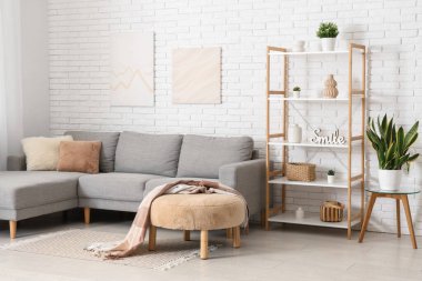 Beyaz tuğla duvarın yanında gri kanepesi ve raf ünitesi olan aydınlık oturma odasının içinde.