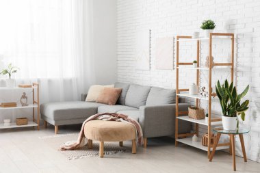 Beyaz tuğla duvarın yanında gri kanepe ve raf üniteleriyle aydınlık oturma odasının içinde.