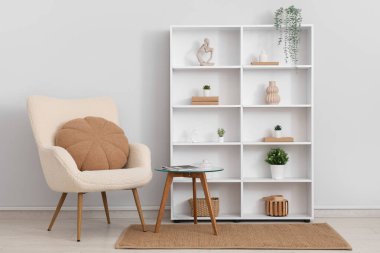 Beyaz duvarın yanında koltuk, masa ve raf ünitesi olan bir oda.