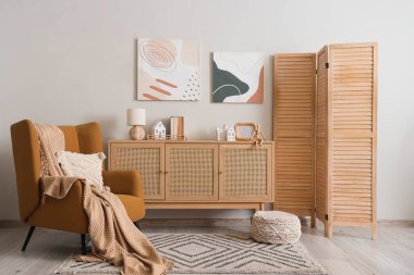 Oturma odasındaki beyaz duvarın yanında ev şekilli mum tutacakları olan çekmeceler ve koltuklar.