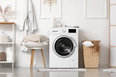 Çamaşır sepetli ve yastıklı modern çamaşır makinesi banyodaki beyaz duvarın yanındaki taburede.