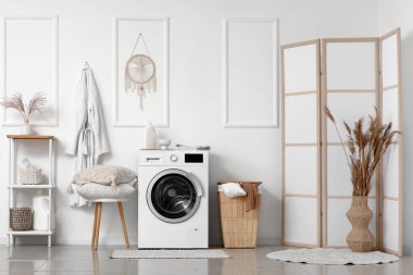 Çamaşır sepetli ve yastıklı modern çamaşır makinesi banyodaki beyaz duvarın yanındaki taburede.