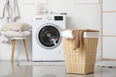 Çamaşır sepetli ve yastıklı modern çamaşır makinesi banyodaki beyaz duvarın yanındaki taburede.