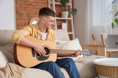 Genç adam evdeki kanepede gitar çalıyor.
