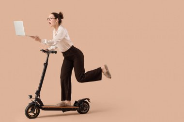 Dizüstü bilgisayarı olan aceleci bir iş kadını bej arka planda elektrikli scooter kullanıyor.