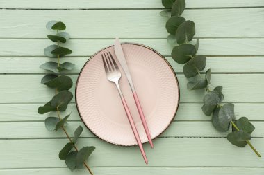 Tabak, çatal bıçak ve okaliptüs dallarıyla yeşil ahşap arka planda şık bir masa.