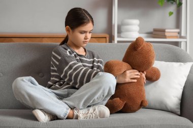 Evdeki kanepede oyuncak ayıyla oturan üzgün kız. Evlat edinme kavramı