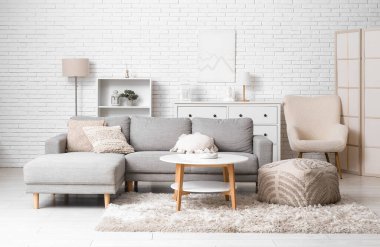 Beyaz tuğla duvarın yanında masa, puf ve kanepe olan ışıklı oturma odasının içinde.