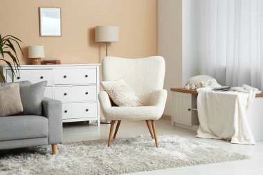 Aydınlık oturma odasının içinde bej duvarın yanında koltuk ve kanepe var.