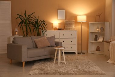 Karanlık oturma odasının içinde çekmeceler, kanepe ve bej duvarın yanında parlayan lambalar var.