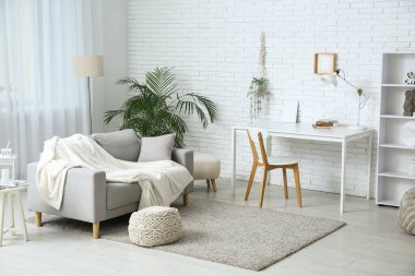 Beyaz tuğla duvarın yanında koltuğu, sandalyesi ve masası olan aydınlık oturma odasının içinde.