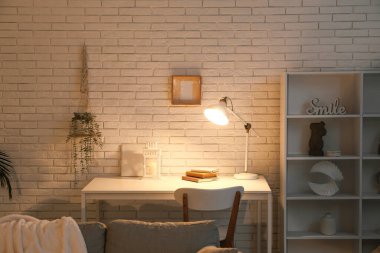 Masası, sandalyesi ve beyaz tuğla duvarın yanında parlayan lambası olan karanlık oturma odasının içinde.