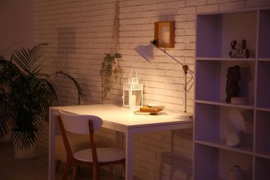 Masası, sandalyesi ve beyaz tuğla duvarın yanında parlayan lambası olan karanlık oturma odasının içinde.