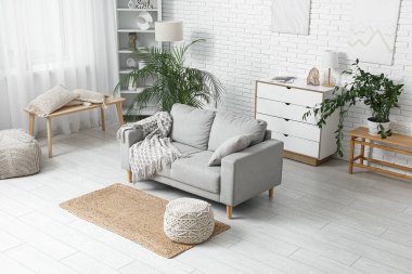 Beyaz tuğla duvarın yanında kanepe, puf ve çiçeklerle aydınlık oturma odasının içinde.