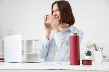 Modern dizüstü bilgisayarlı genç bir iş kadını çalışma odasında çay içiyor, açık termosa odaklanıyor.