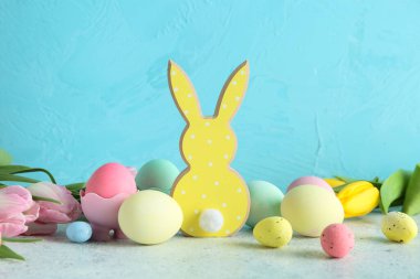 Çiçekler, Paskalya yumurtaları ve mavi duvarın yanındaki gri masada ahşap tavşanlar.