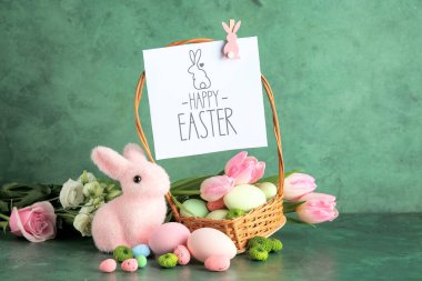 Hasır sepetli kompozisyon, Paskalya yumurtaları, dekoratif tavşancık, yazılı tebrik kartı Mutlu Paskalyalar ve yeşil grunge arka planda çiçekler