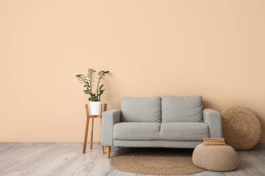 Oturma odasının iç tarafındaki bej duvarın yanında puf, saksı ve gri kanepe.