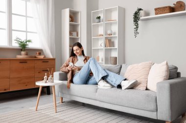 Genç bir kadın kanepede uzanıyor ve oturma odasında cep telefonu kullanıyor.