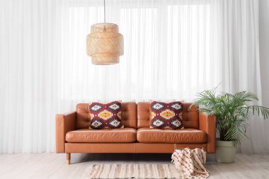 Oturma odasının içinde kahverengi kanepe, palmiye ağacı ve penceresi var.