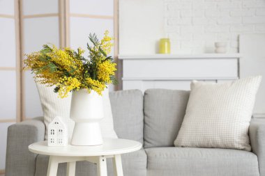 Mimoza çiçekli vazo ve oturma odasındaki masada duran ev figürü.