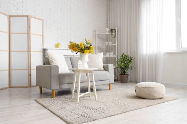 Oturma odasının içinde kanepe, raf ünitesi ve vazoda mimoza çiçekleri var.