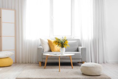 Oturma odasının içinde kanepe, masa ve vazoda mimoza çiçekleri var.