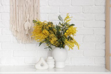 Beyaz tuğla duvarın yanındaki rafta mimoza çiçekleri olan vazo.