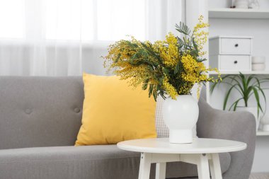 Oturma odasındaki kanepenin yanındaki masada mimoza çiçekleri olan vazo.