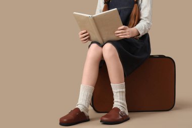 Takunyalı genç kız, bej arka planda bavulla ilgili kitap okuyor.