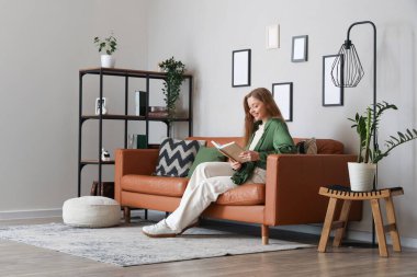 Genç bir kadın kanepede oturuyor ve oturma odasında kitap okuyor.