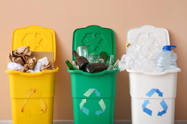 Geri dönüşüm sembollü çöp kutuları ve bej duvarın yanındaki farklı çöpler.