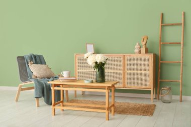 Kahve masasında beyaz şakayık vazosu, sandalye ve şifonyer yeşil duvarın yanında.