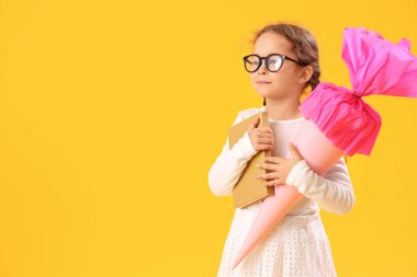 Pembe okul konisi ve sarı arka plan kitabı olan gözlüklü mutlu küçük kız.