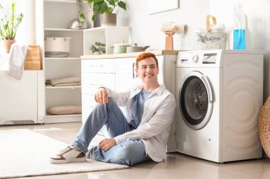 Genç adam evde çamaşır makinesinin yanında oturuyor.