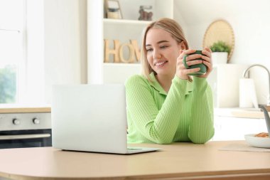 Bir fincan sıcak çay ve dizüstü bilgisayarlı genç bir kadın mutfaktaki masada oturuyor.