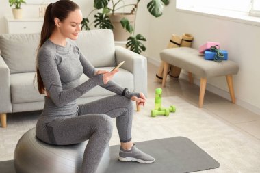 Sportif genç bayan evde fitball oynarken cep telefonu kullanıyor.