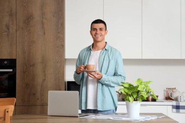 Masasında modern dizüstü bilgisayarı olan mutfakta kahve içen yakışıklı bir genç.