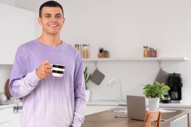 Masasında modern dizüstü bilgisayarı olan mutfakta kahve içen yakışıklı bir genç.